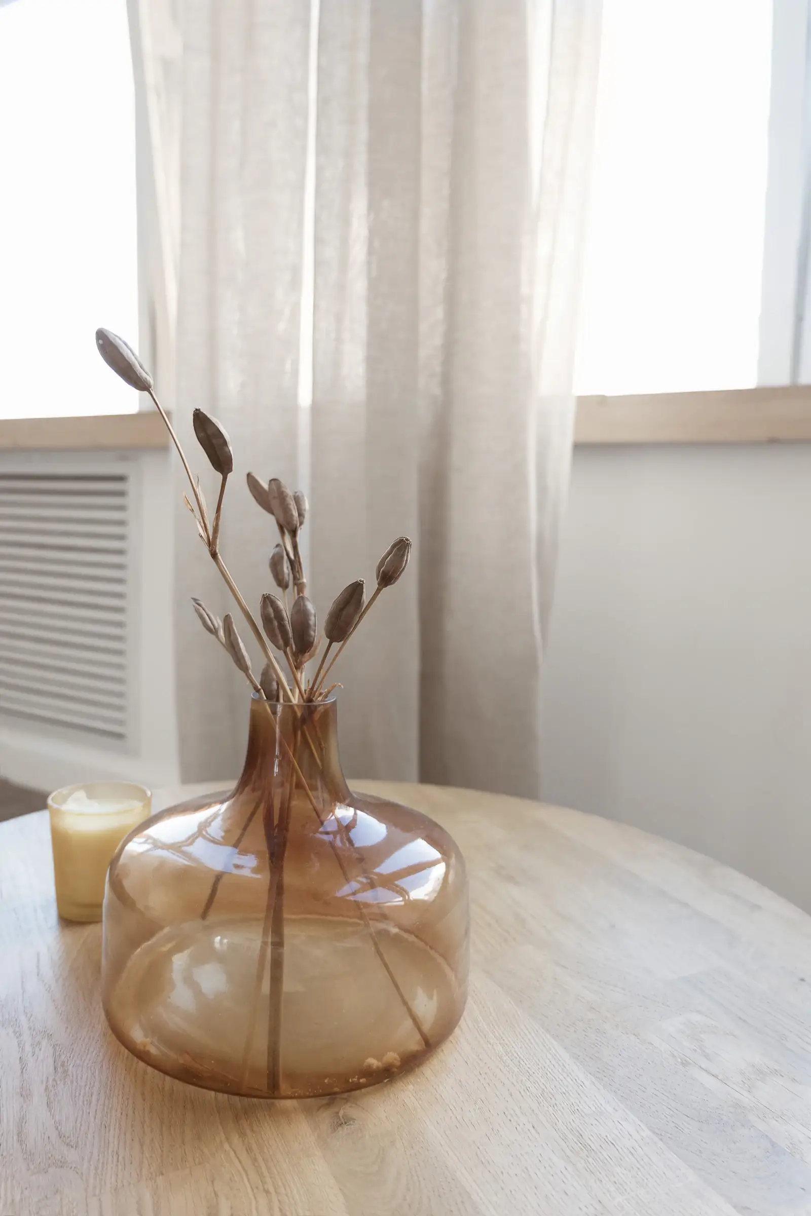 Amber glass vase with dried stems and a candle on a pale wooden table by a linen curtain